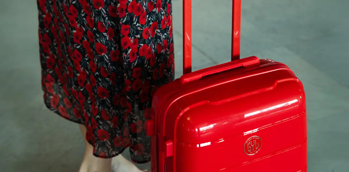 Femme en robe à motifs floraux tenant une valise cabine GYL rigide rouge, dans un espace intérieur lumineux – bagage compact et coloré au style affirmé.
