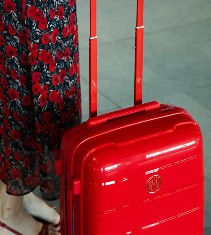 Femme en robe à motifs floraux tenant une valise cabine GYL rigide rouge, dans un espace intérieur lumineux – bagage compact et coloré au style affirmé.