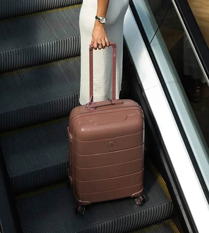 Femme en robe blanche montant un escalator avec une valise rigide GYL marron, sous un éclairage tamisé – bagage de soute élégant et durable.
