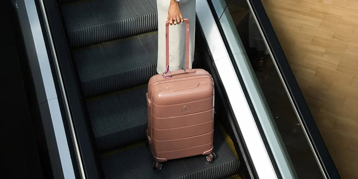 Valise rigide GYL Citadine en polycarbonate rose poudré, tirée sur un escalator, idéale pour les voyages urbains et pratiques.