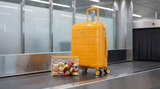 Une valise cabine rigide de couleur jaune vif posée sur un tapis roulant de contrôle de sécurité aéroportuaire, à côté d'une pochette transparente remplie de macarons colorés et de fruits frais (fraises, framboises, myrtilles).