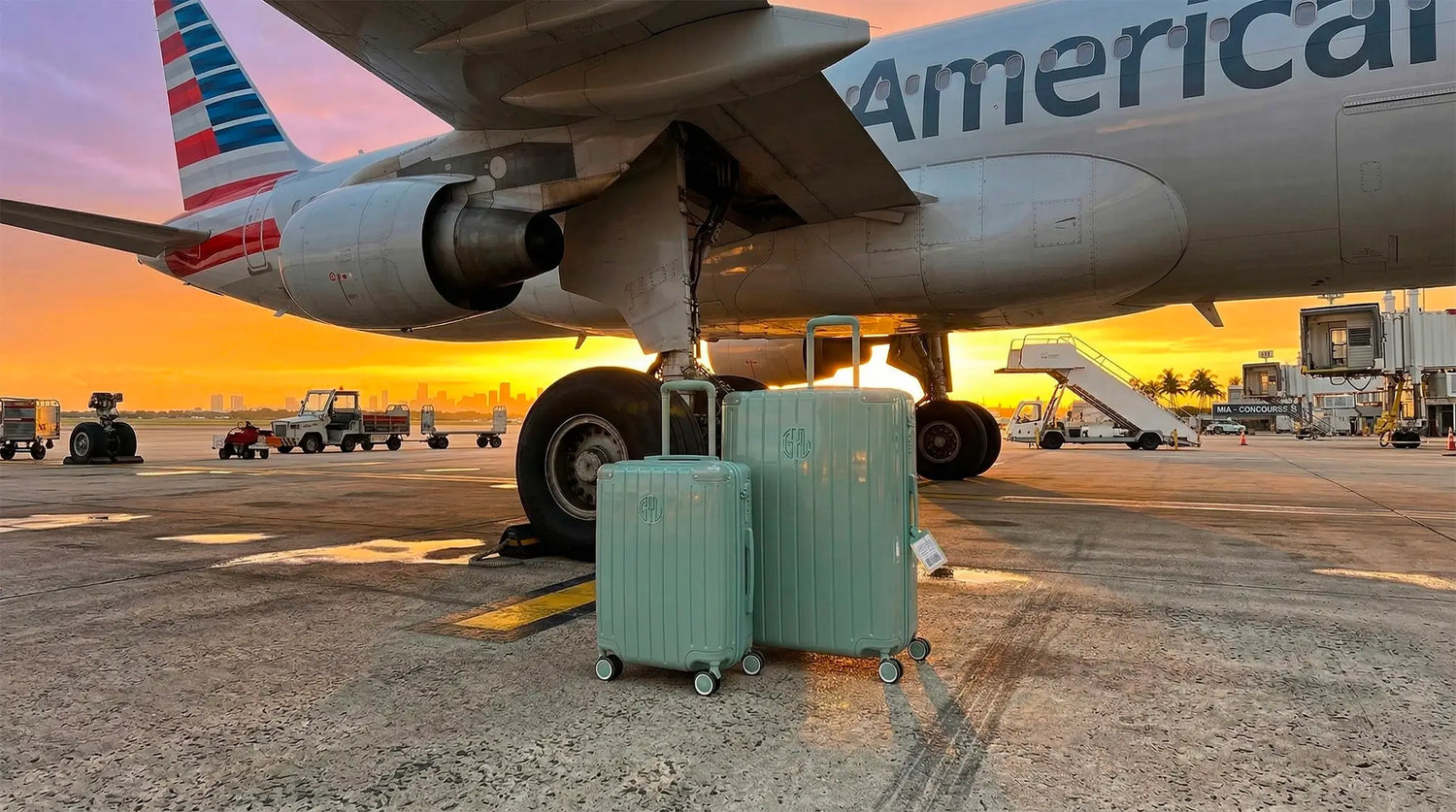 Set de deux bagages GYL vert pastel posés au sol sur une piste d'aéroport, situés sous le réacteur d'un avion de ligne American Airlines avec un ciel orangé de coucher de soleil en arrière-plan.
