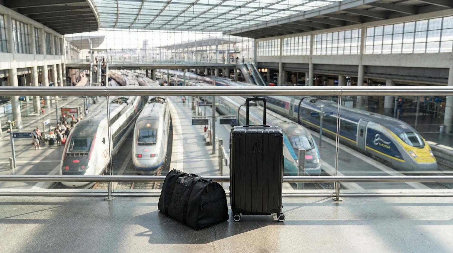 Valise rigide GYL et sac de voyage GYL posés au-dessus des quais d’une grande gare, avec plusieurs trains grande vitesse en arrière-plan, illustrant les règles de bagages dans le train