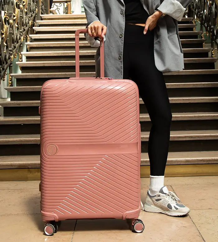 Vue en contre-plongée d’une valise rigide GYL rose poudré devant un grand escalier métallique, tirée par une femme en blazer gris – bagage grand format haut de gamme.