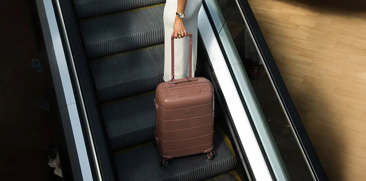 Femme en robe blanche montant un escalator avec une valise rigide GYL marron, sous un éclairage tamisé – bagage de soute élégant et durable.