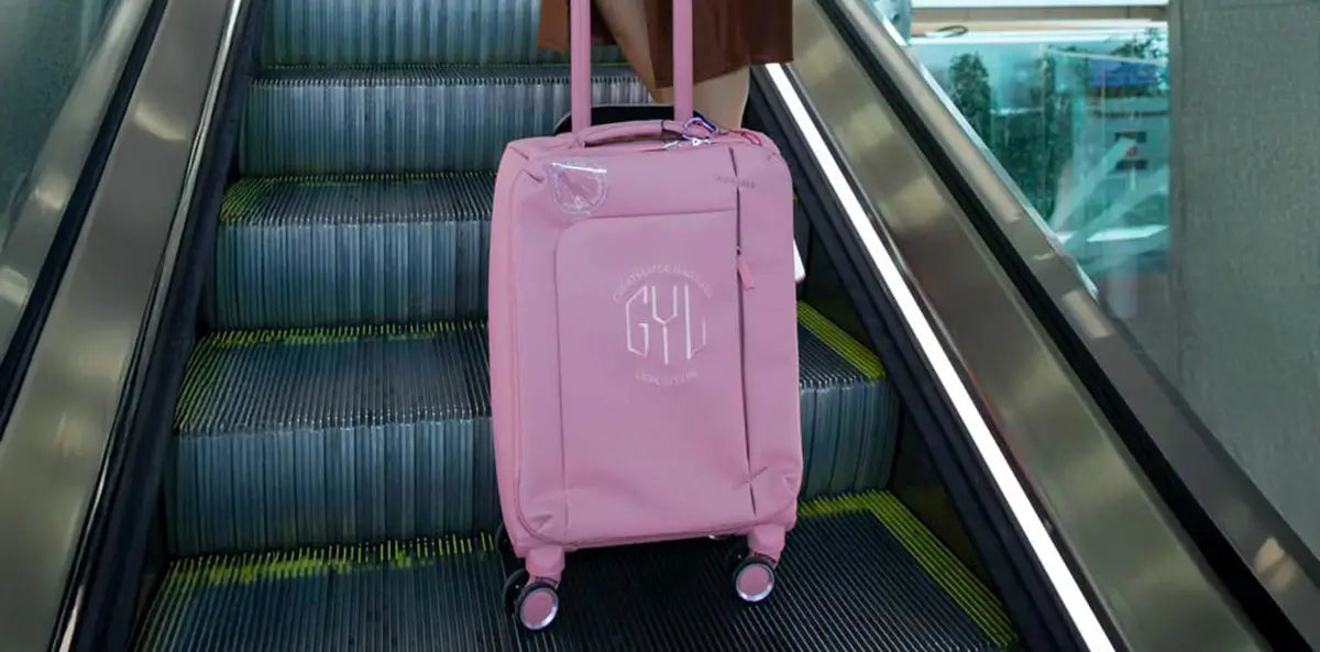 Valise souple GYL rose poussée par une femme en jupe marron, vue en gros plan sur un escalator d’aéroport – bagage léger idéal pour les courts séjours.