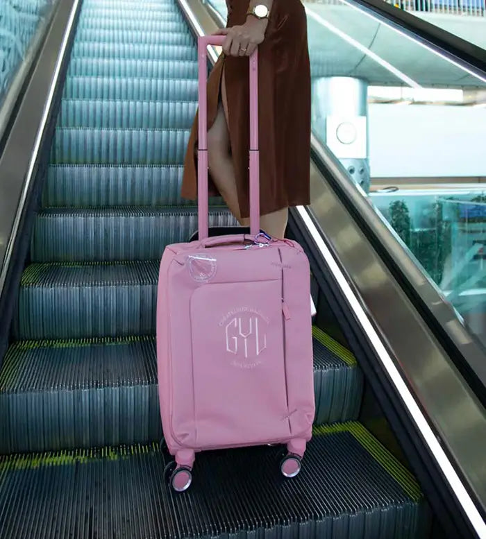 Valise souple GYL rose poussée par une femme en jupe marron, vue en gros plan sur un escalator d’aéroport – bagage léger idéal pour les courts séjours.