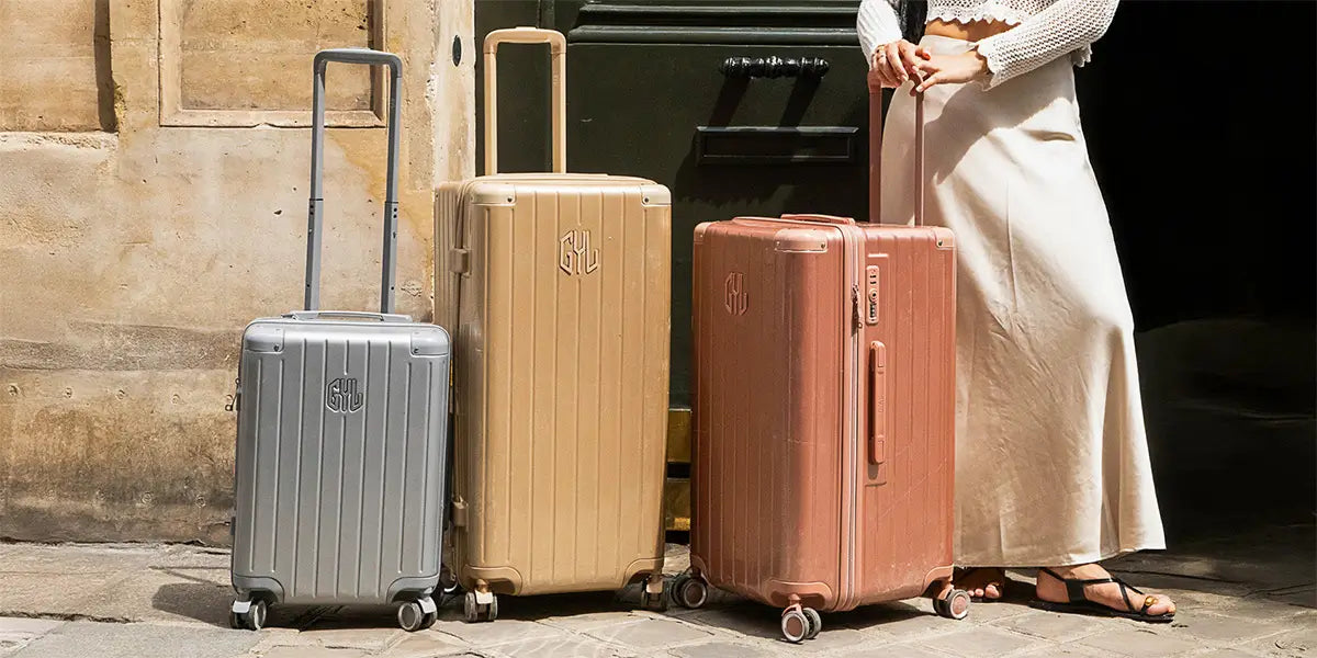 Trois valises rigides GYL de la collection TRUNK en aluminium, couleurs argent, doré et rose, posées devant un mur en pierre avec une personne tenant la grande valise.