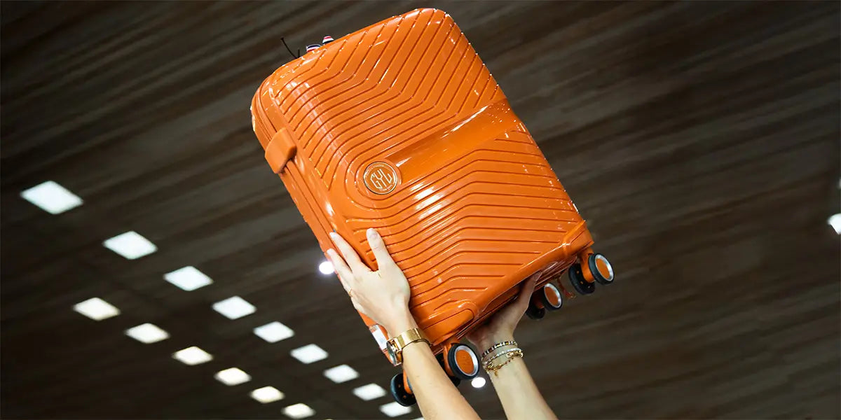 Valise rigide GYL Signature en polycarbonate orange, tenue en l’air avec ses quatre roulettes visibles, design moderne et robuste.