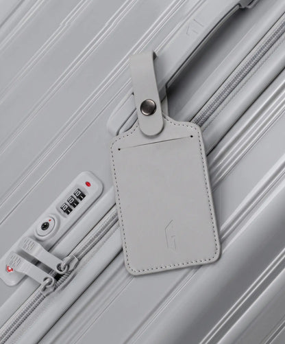 Etiquette de bagage GYL en cuir coloris gris argent fixee par sa bride a pression sur la poignee d'une valise GYL assortie