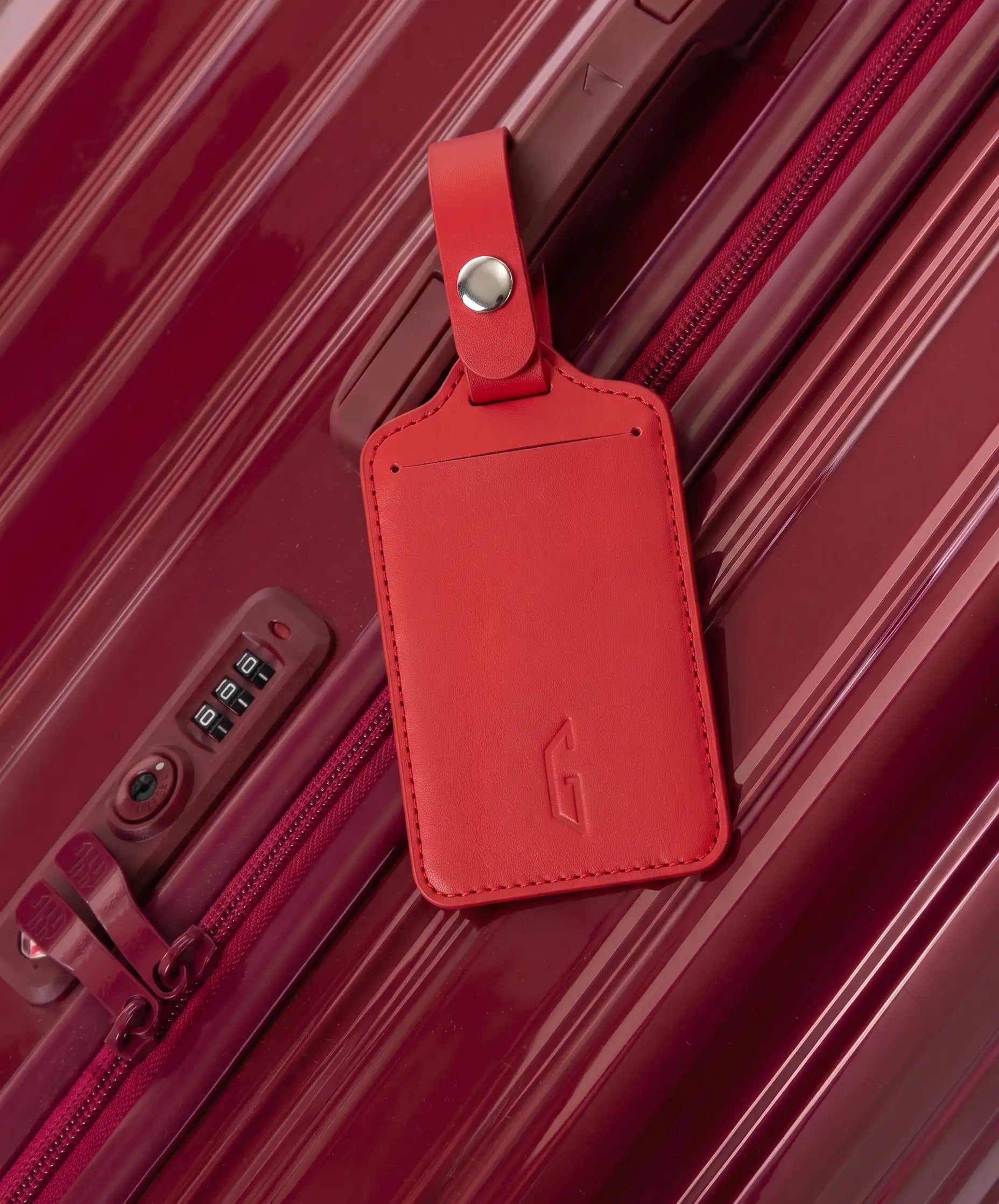Etiquette de bagage GYL en cuir coloris rouge fonce fixee par sa bride a pression sur la poignee d'une valise GYL assortie