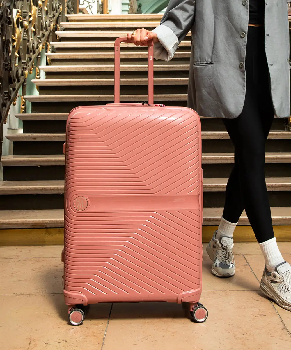 Gros plan sur une valise rigide GYL grand format rose, posée devant un escalier ancien, tenue par une femme en veste grise – bagage haut de gamme au style affirmé.