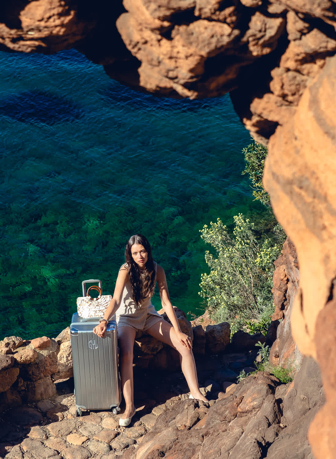 Femme avec valise trunk GYL grise dans une crique rocheuse au bord de la mer turquoise