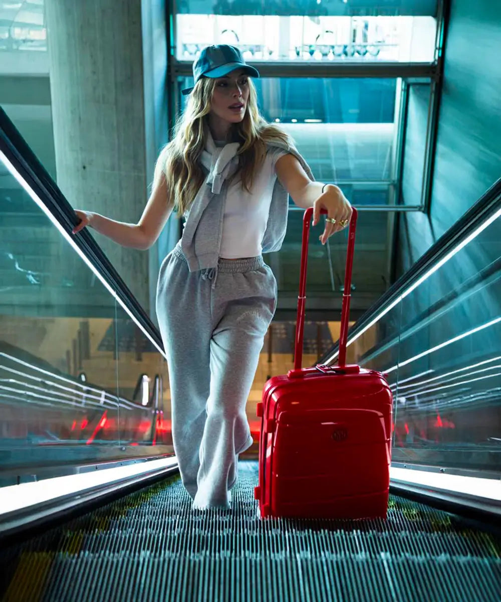 Jeune femme sportive descendant un escalator avec une valise cabine rigide GYL rouge, habillée en survêtement gris clair – bagage compact et stylé pour les voyageurs dynamiques.