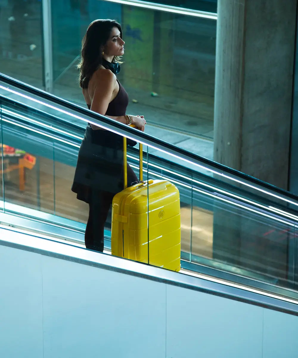 Femme chic montant sur un escalator avec une valise rigide GYL jaune, dans un aéroport moderne – bagage coloré au design premium, idéal pour les city breaks.