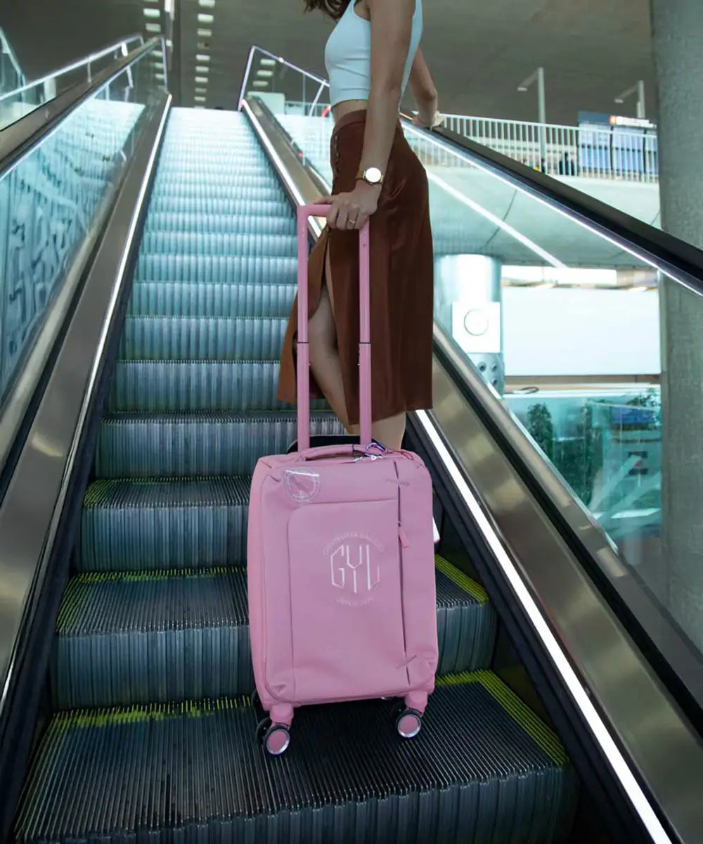 Femme en jupe longue montant un escalator avec une valise souple GYL rose, dans un terminal d’aéroport – bagage léger et féminin, parfait pour les déplacements rapides.