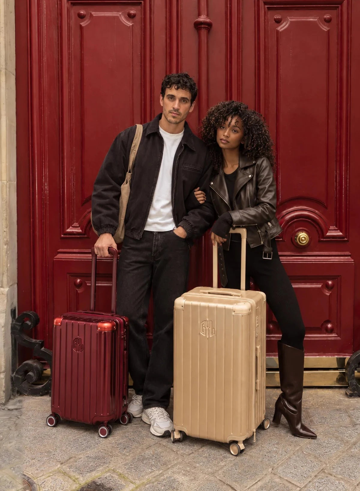 Couple posant devant une porte cochère rouge à Paris avec deux valises rigides GYL dark red et champagne